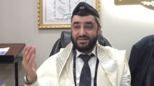 Bearded man wearing ceremonial white robes and a kippah, seated in an office chair, gesturing with his right hand as he speaks.