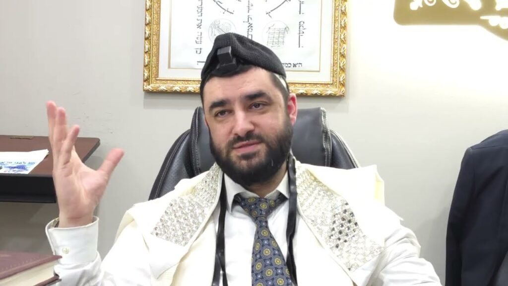 Rabbi in a white prayer shawl and business attire sits in an office chair, gesturing with his left hand.