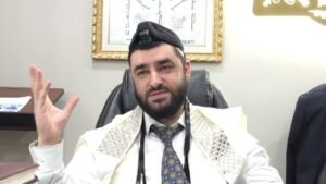 Rabbi in a white prayer shawl and business attire sits in an office chair, gesturing with his left hand.