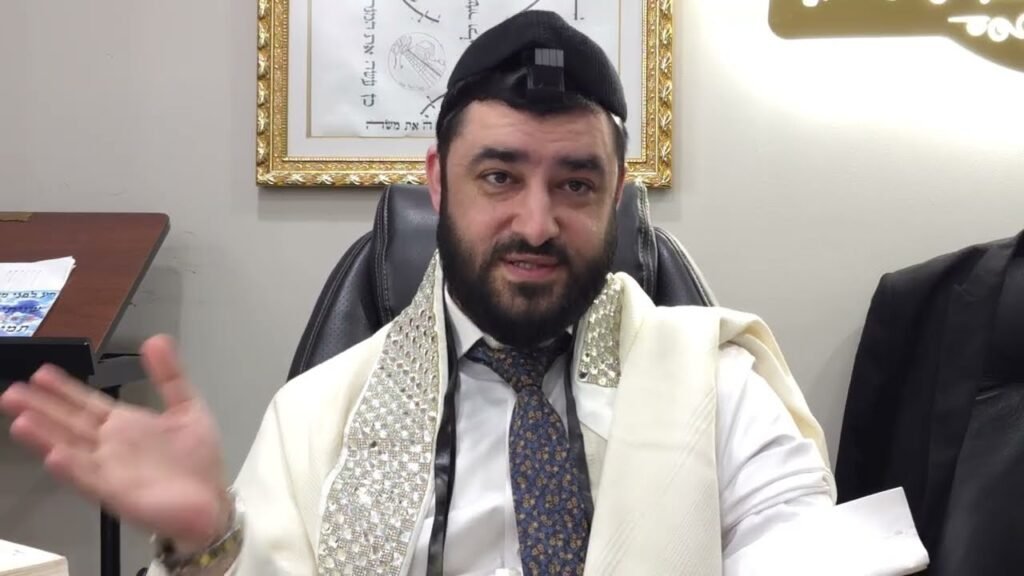 A bearded man wearing a white ceremonial robe and decorative shawl sits at a desk, gesturing with his left hand while speaking in an office setting, with framed artwork in the background.
