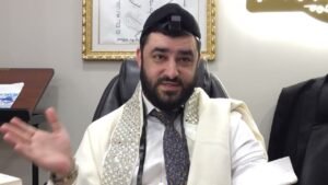 A bearded man wearing a white ceremonial robe and decorative shawl sits at a desk, gesturing with his left hand while speaking in an office setting, with framed artwork in the background.