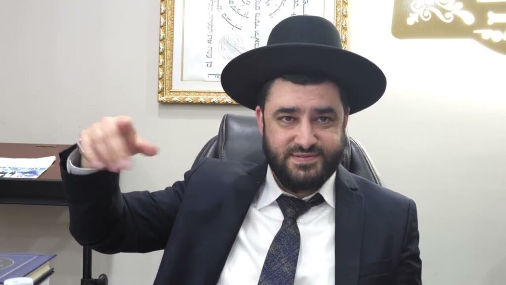 Bearded man in a dark suit and black hat sits at a desk, gesturing with his right hand toward the camera.