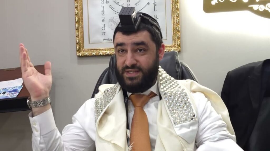 Bearded man in a white shirt and orange tie, wearing tefillin and a jeweled shawl, gesturing with his left hand.