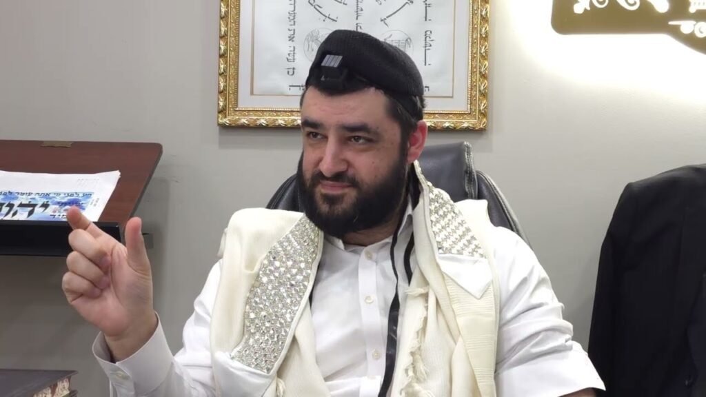 Bearded man in white traditional robes sitting in an office, wearing a small camera on a black cap and gesturing with his finger.