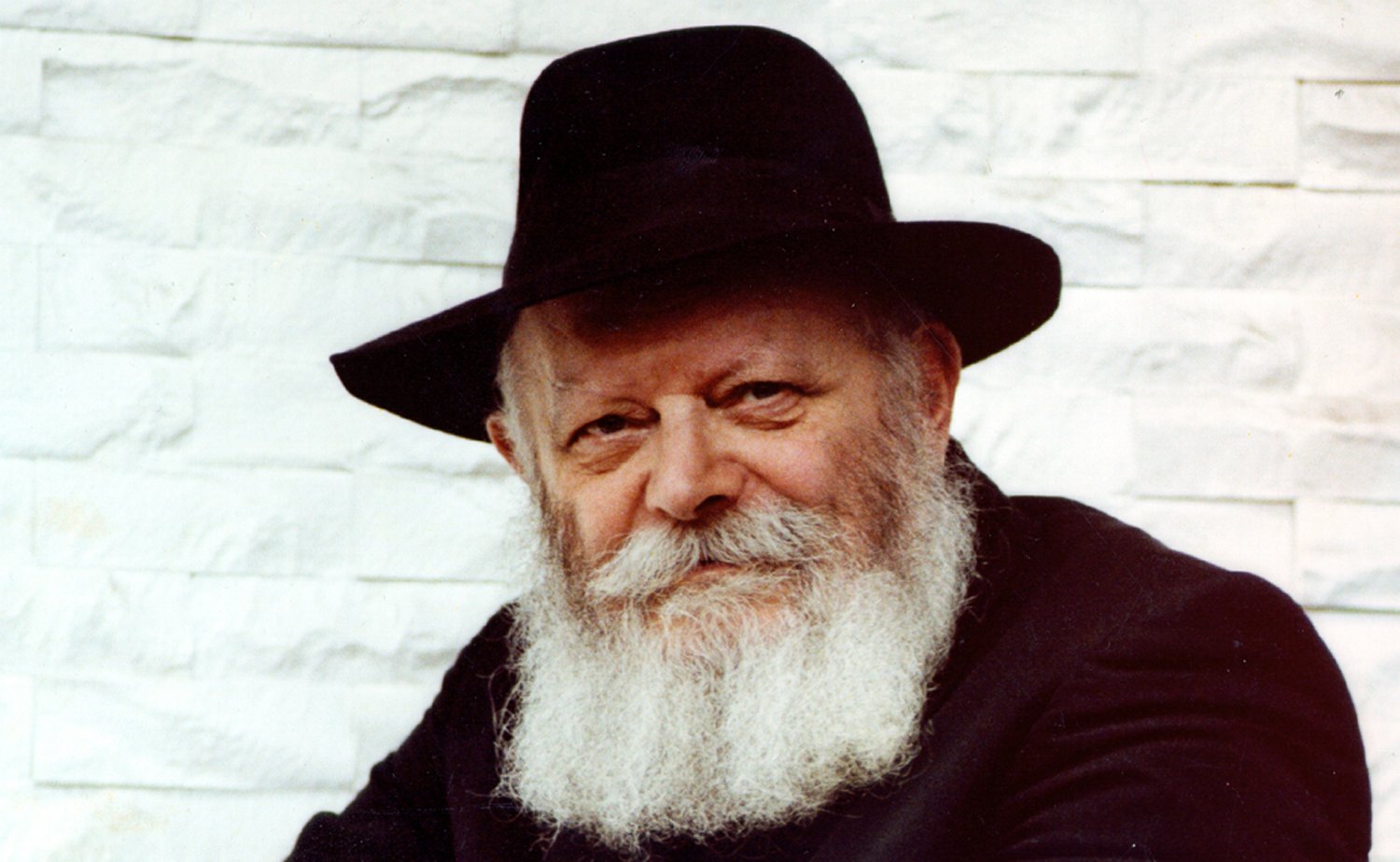 Portrait of a bearded older man in a wide-brimmed black hat, standing against a white brick wall.
