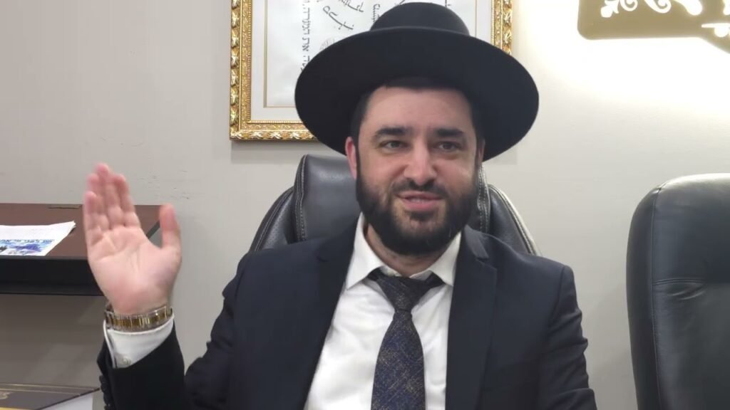 Bearded man in a suit and hat sits at a desk and raises his right hand in greeting in an office setting.