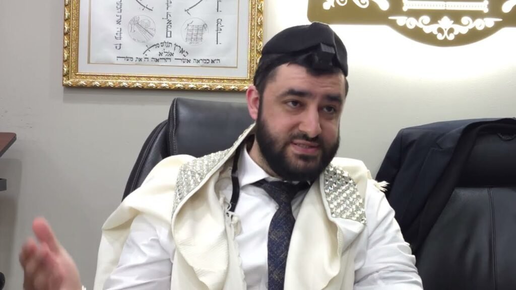 Bearded man wearing a white tallit with silver embellishments and a kippah, seated in an office, gesturing while speaking.