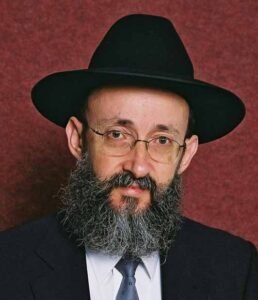 Man with a black fedora, glasses, and full beard looks at the camera against a reddish backdrop, wearing a dark suit and tie.