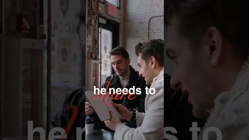 Two men sit at a cafe counter, looking at an open book together; a third person is visible on the right.