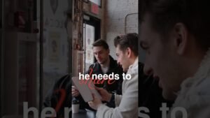 Two men sit at a cafe counter, looking at an open book together; a third person is visible on the right.