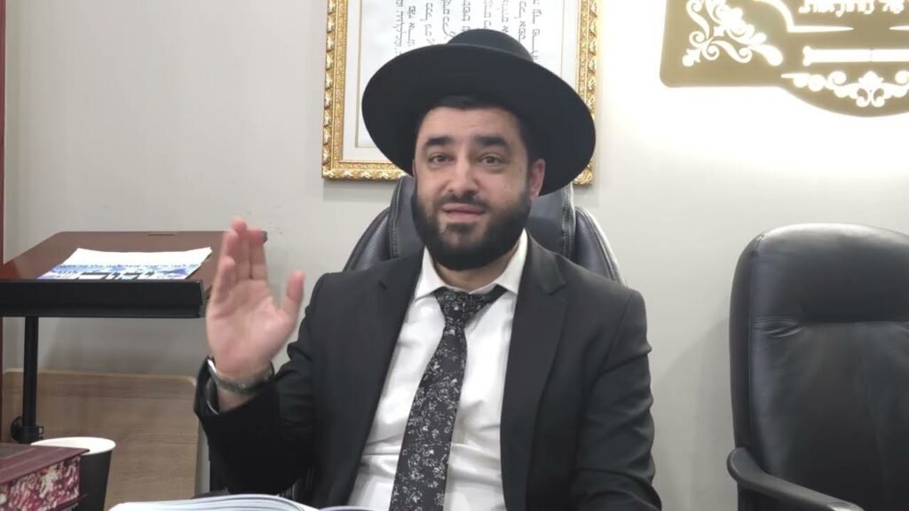 Bearded man in a black suit and wide-brimmed hat sits at a desk in an office, raising his right hand as he speaks.