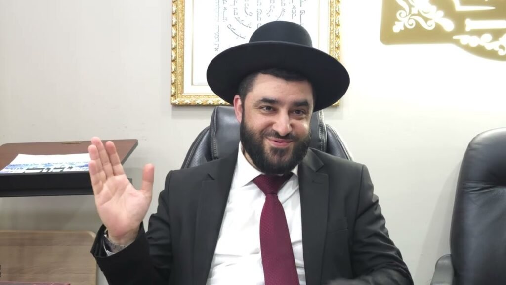 Bearded man in a black suit and wide-brim hat sits at a desk, waving hello.