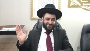 Bearded man in a black suit and wide-brim hat sits at a desk, waving hello.