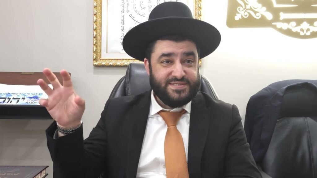 Bearded man in a black suit and orange tie sits at a desk wearing a wide-brimmed black hat, gesturing with his left hand as he speaks.