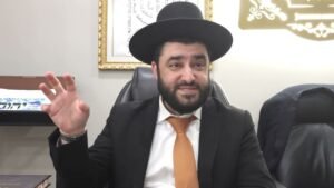 Bearded man in a black suit and orange tie sits at a desk wearing a wide-brimmed black hat, gesturing with his left hand as he speaks.