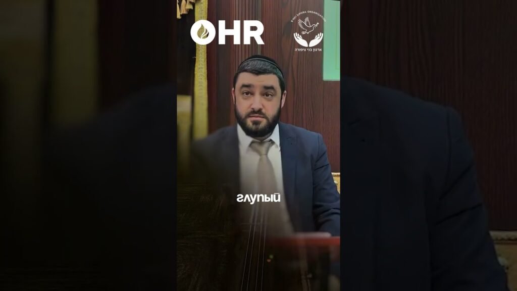 Bearded man in a suit seated at a podium during a formal event, with HR logo and an organizational emblem in the background.”