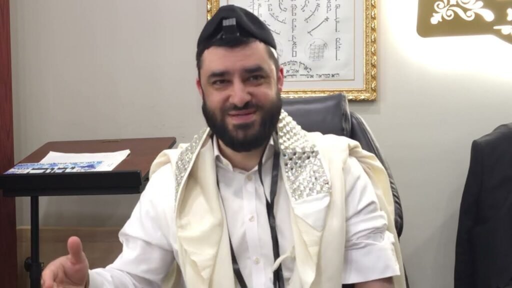 Bearded man in a white shirt and cream ceremonial robe with sparkling shoulder decorations, seated in an office with a desk nearby and framed art behind him (kippah on head).