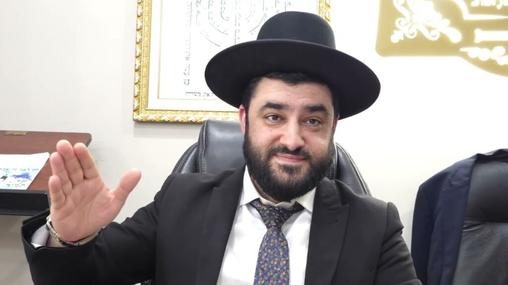 Bearded man in a black suit and wide-brimmed hat sits at a desk in an office, waving with his right hand, smiling at the camera, framed artwork behind him
