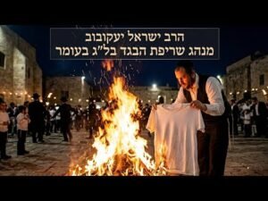 Man in a vest and white shirt holds a folded shirt over a large bonfire in a nighttime courtyard, as onlookers gather and a Hebrew banner hangs above.