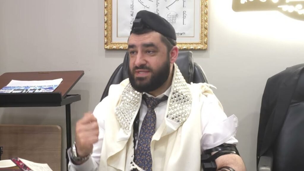 Bearded man wearing a kippah and ornate scarf speaks in an office setting.