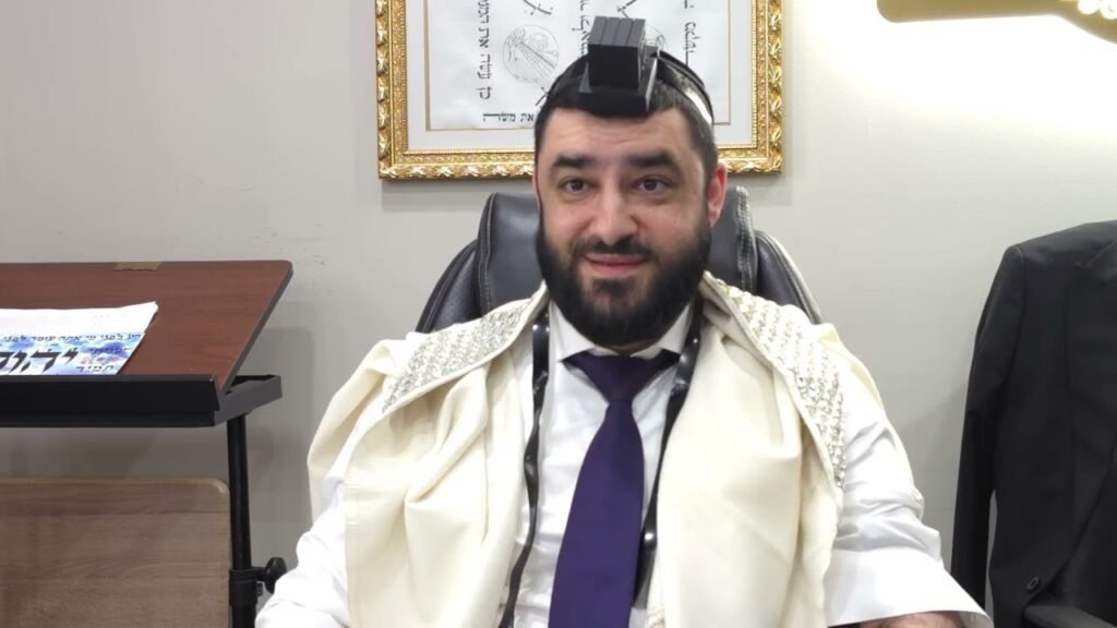 Bearded man wearing a tallit and tefillin sits in a conference chair in an office, looking at the camera.