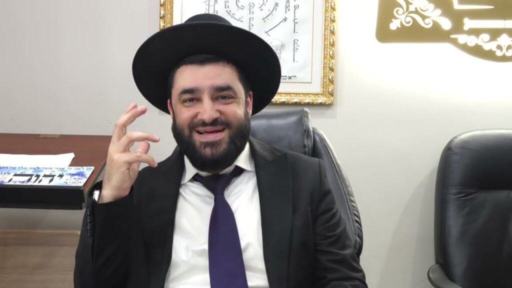 Smiling bearded man in a black hat and suit with a purple tie, seated in a leather office chair, making a hand gesture; framed artwork on the wall behind and another empty chair to the right.