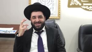 Smiling bearded man in a black hat and suit with a purple tie, seated in a leather office chair, making a hand gesture; framed artwork on the wall behind and another empty chair to the right.