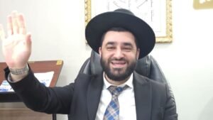 Man in a dark suit and wide-brimmed hat sits at a desk in an office, smiling and waving at the camera.
