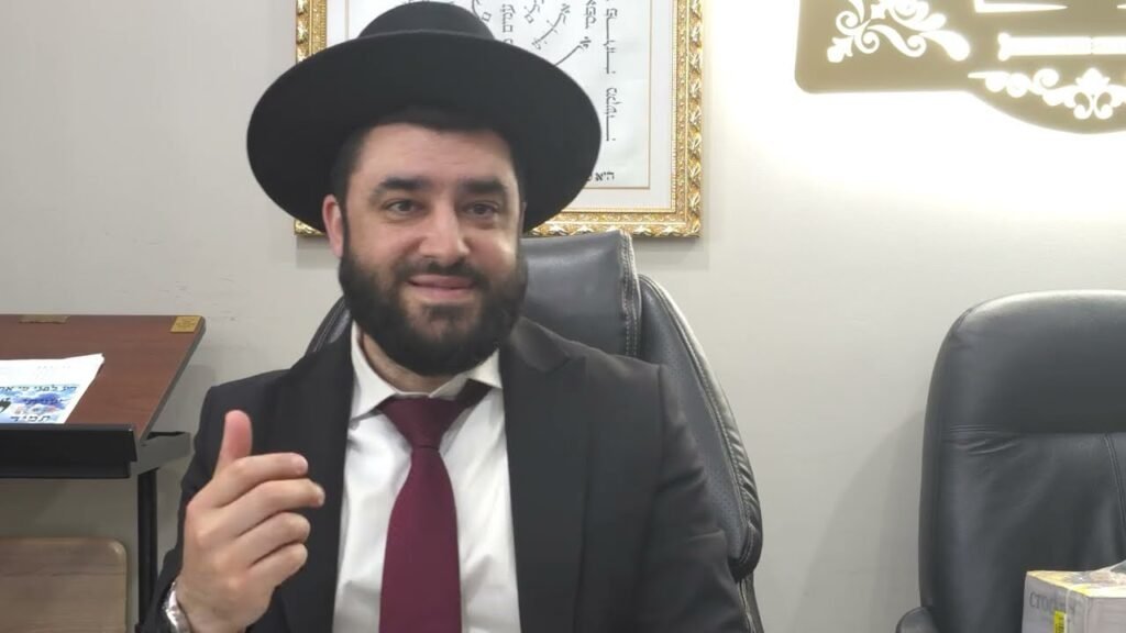 Bearded man in a black suit and hat sits at a desk, gesturing with his right hand in an office setting.