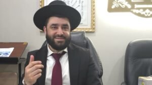 Bearded man in a black suit and hat sits at a desk, gesturing with his right hand in an office setting.
