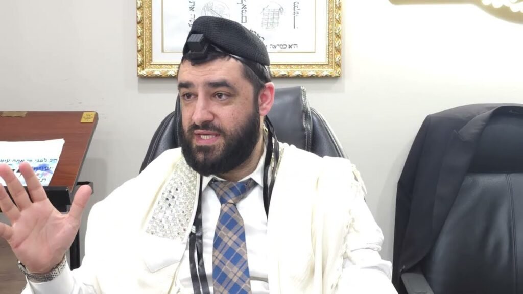 Rabbi with tefillin on his head and cream prayer shawl gesturing while speaking from an office chair.