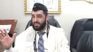 Rabbi with tefillin on his head and cream prayer shawl gesturing while speaking from an office chair.