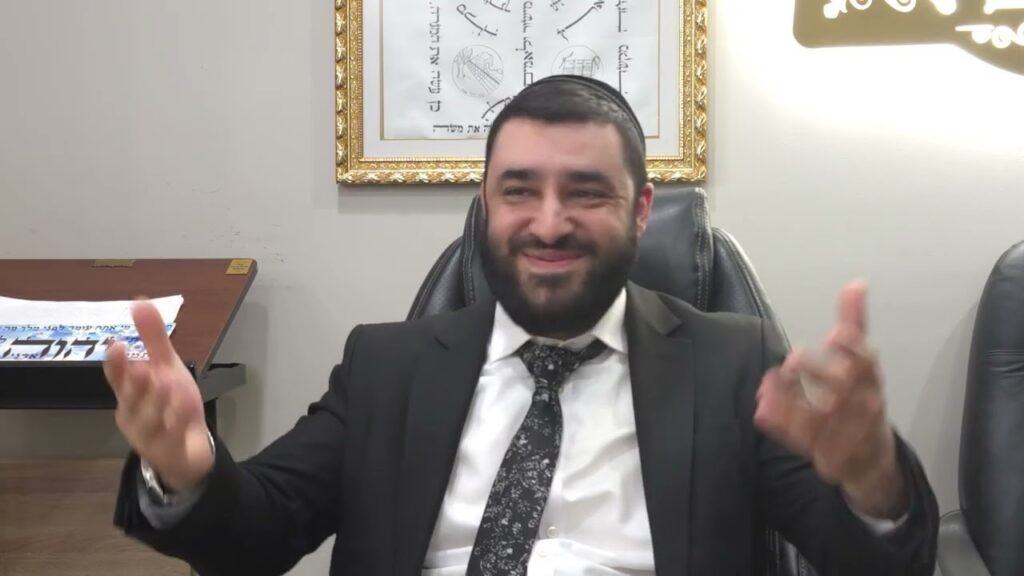 Smiling man in a suit sits in a leather chair, hands raised in conversation in an office setting with a framed artwork behind him.