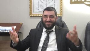 Smiling man in a suit sits in a leather chair, hands raised in conversation in an office setting with a framed artwork behind him.
