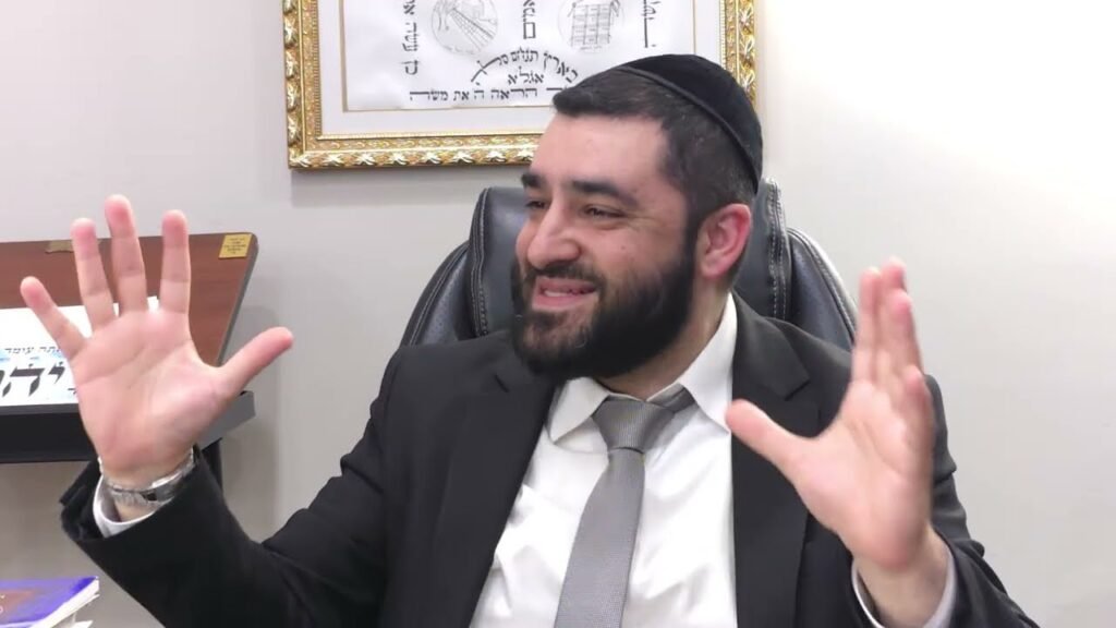 Man wearing a suit and kippah sits at a desk, smiling, with hands raised as if explaining something in a meeting room.