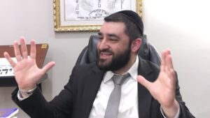 Man wearing a suit and kippah sits at a desk, smiling, with hands raised as if explaining something in a meeting room.