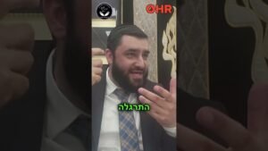 Bearded man in a suit speaking and gesturing with his hands, seated indoors; logos visible at the top (OHR) and a circular emblem in the background, with green Hebrew text across the bottom.