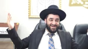 Smiling bearded man in a black hat and suit, arms raised, seated in an office chair during a meeting.