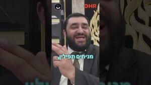 Smiling man in a suit in a formal setting, with decorative gold backdrop and OHR logo in the corner and Hebrew subtitle text overlay.