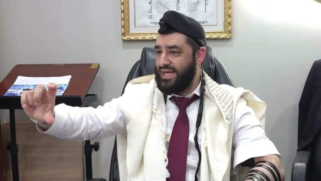 Bearded man in a white robe with a red tie sits at a desk, gesturing with his left hand while speaking in an office setting.