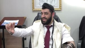 Bearded man in a white robe with a red tie sits at a desk, gesturing with his left hand while speaking in an office setting.