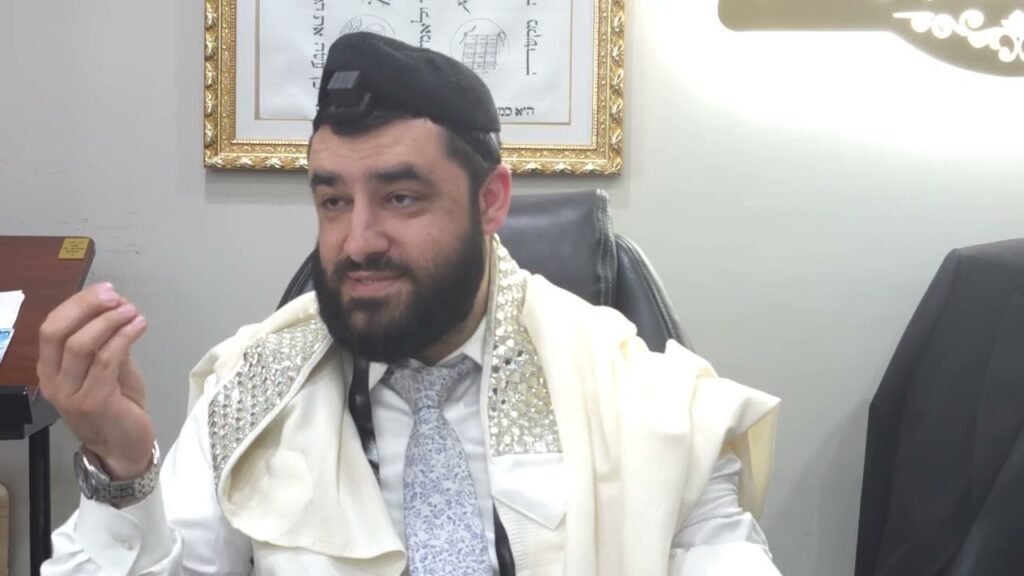Bearded man wearing a prayer shawl and kippah sits at a desk, gesturing with his hand during a discussion.