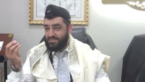 Bearded man wearing a prayer shawl and kippah sits at a desk, gesturing with his hand during a discussion.