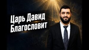 Man in a dark suit with a white shirt and patterned tie stands to the right, in front of a backdrop with large white Cyrillic text that reads “Царь Давид Благословит.”