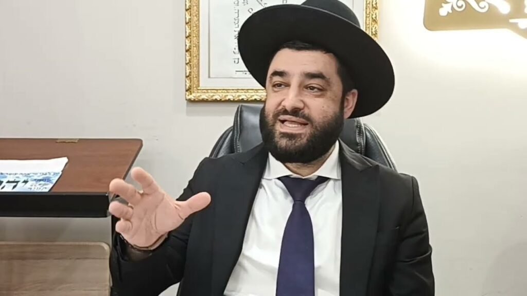 Bearded man in a black suit and tie wearing a wide-brimmed hat, seated at a desk and gesturing with his hand.