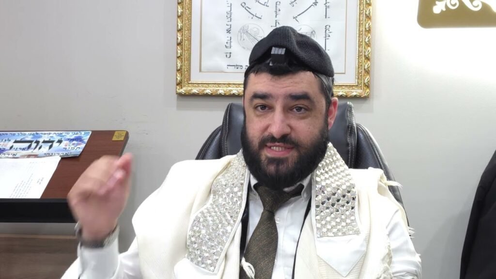 Rabbi with a beard wearing a kippah and prayer shawl sits in a chair in an office.