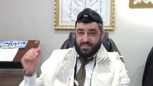 Rabbi with a beard wearing a kippah and prayer shawl sits in a chair in an office.