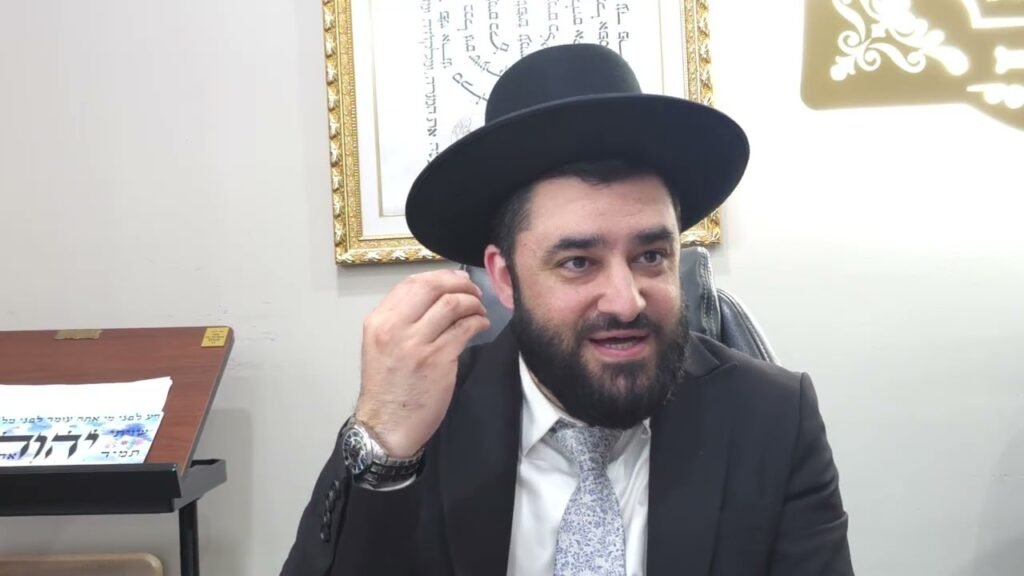 Bearded man in a suit and black hat adjusts his ear while seated at a desk in an office setting.