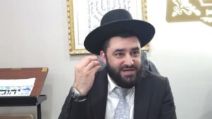 Bearded man in a suit and black hat adjusts his ear while seated at a desk in an office setting.