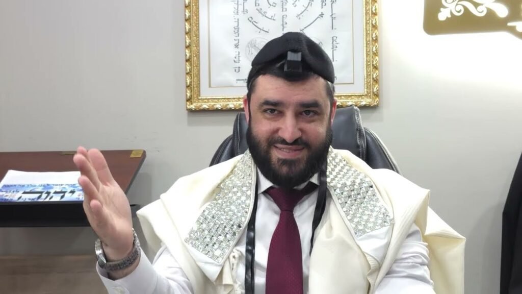 Bearded man in ceremonial white robe with silver-embroidered shawl and burgundy tie sits at a desk, hand raised in greeting.